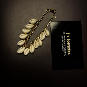 Un bracelet à maillons dorés orné d'une cascade de coquillages cauris blancs disposé de manière longiligne à côté d'une carte noire jekouin.