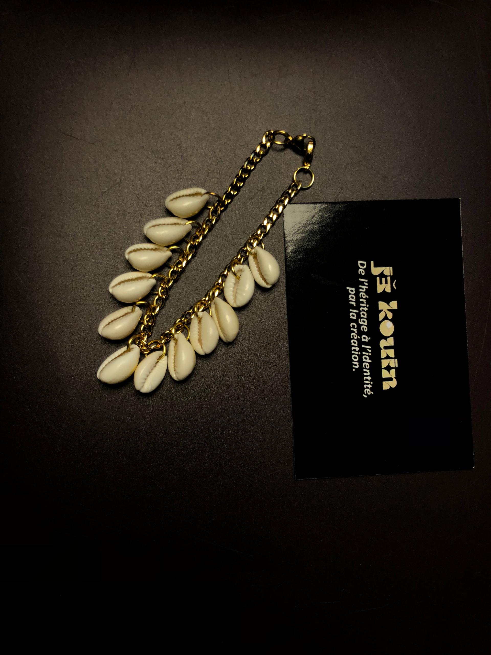 Un bracelet à maillons dorés orné d'une cascade de coquillages cauris blancs disposé de manière longiligne à côté d'une carte noire jekouin.