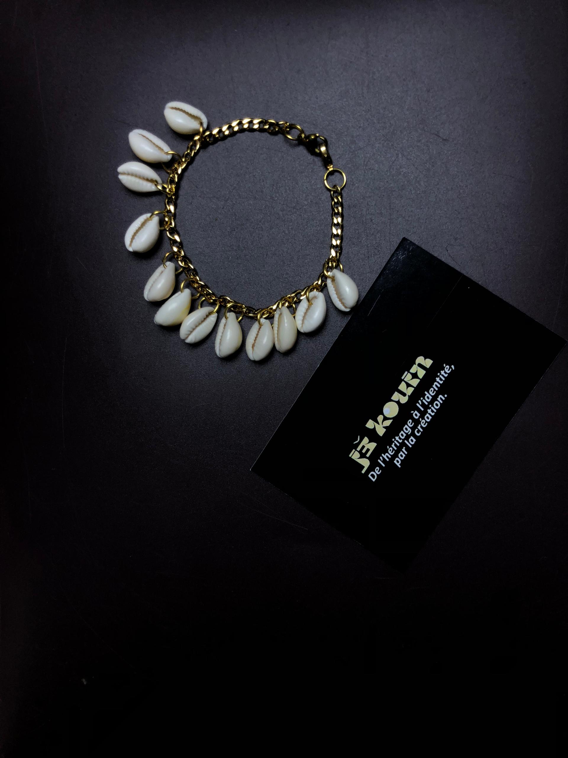 Vue de dessus d'un bracelet en chaîne dorée et cauris disposé en cercle à côté de la carte noire jekouin sur fond sombre.