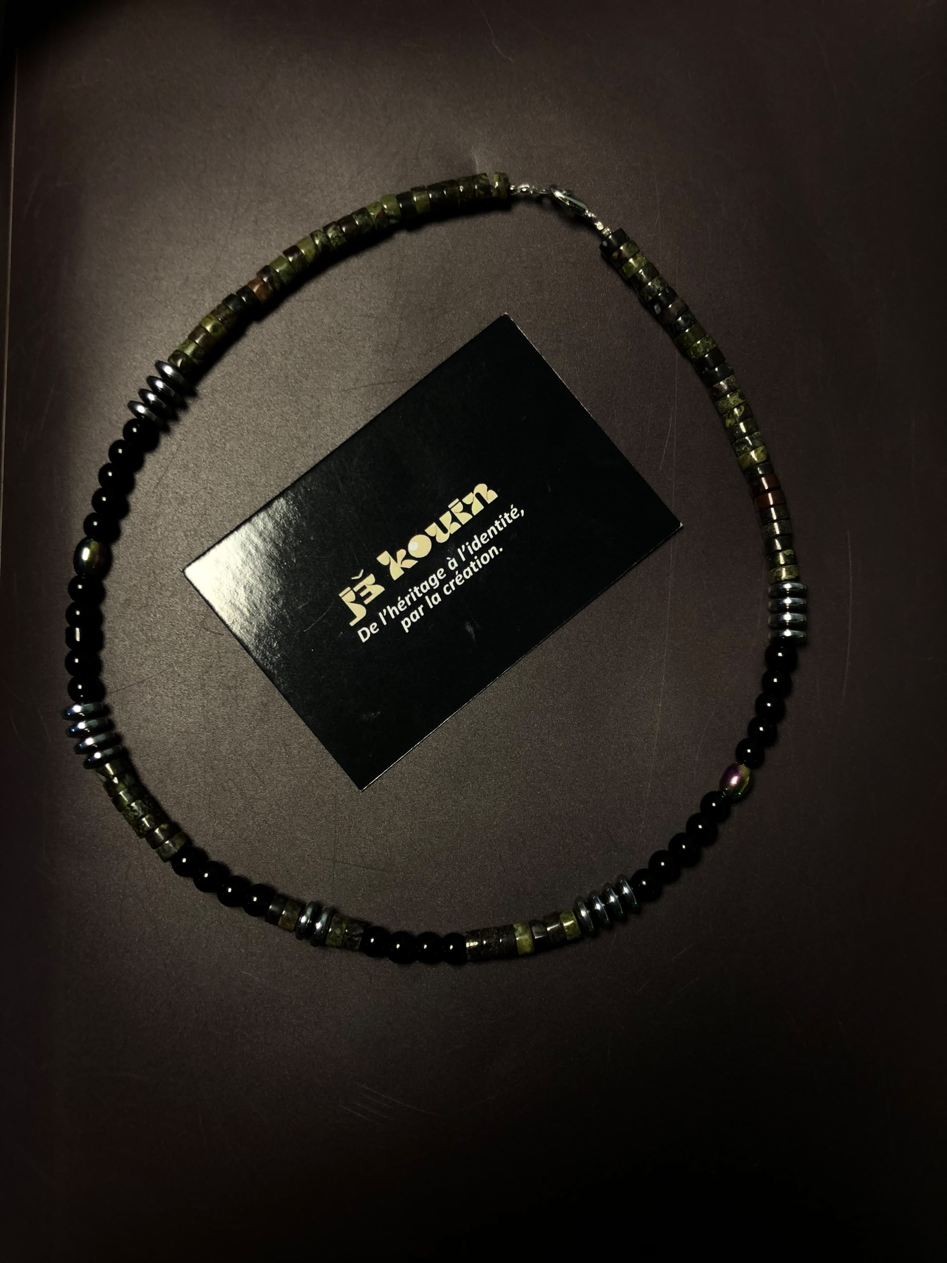 Collier ras-de-cou mixte composé de perles cylindriques vertes, rondes noires et hématite métallisée, entourant la carte jekouin sur fond sombre.