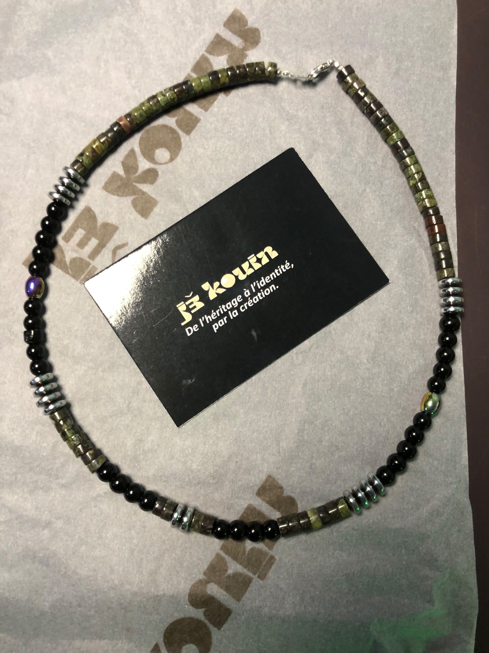 Vue de dessus du collier unisexe jekouin posé sur du papier de soie imprimé au nom de la marque avec sa carte de visite au centre.