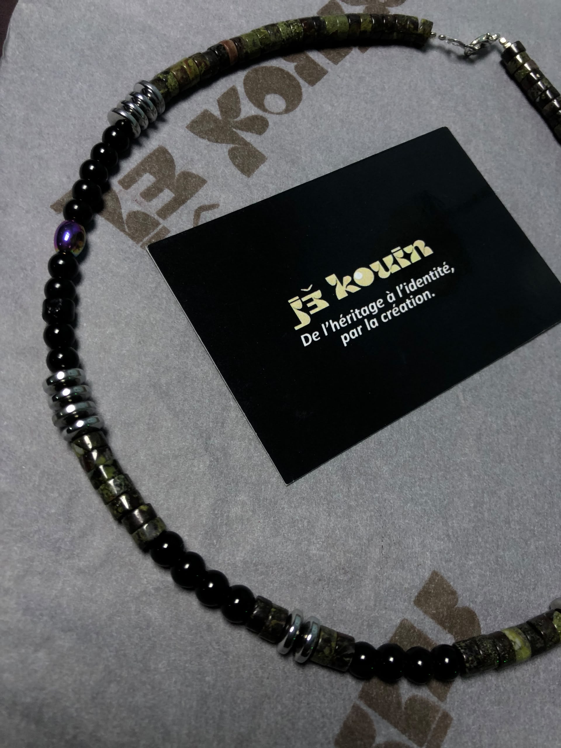 Plan rapproché montrant le détail des perles cylindriques vertes, des rondelles d'hématite et de la perle irisée du collier jekouin.