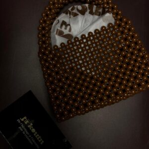 Sac à main artisanal tissé en grosses perles marron chocolat avec anses intégrées et doublure en tissu imprimé jǯ kouin, posé à côté d'une carte de visite sur fond sombre.