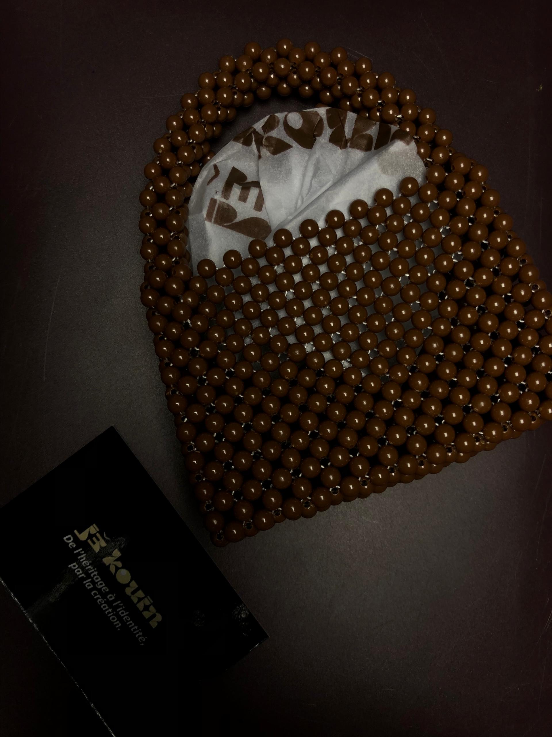 Sac à main artisanal tissé en grosses perles marron chocolat avec anses intégrées et doublure en tissu imprimé jǯ kouin, posé à côté d'une carte de visite sur fond sombre.