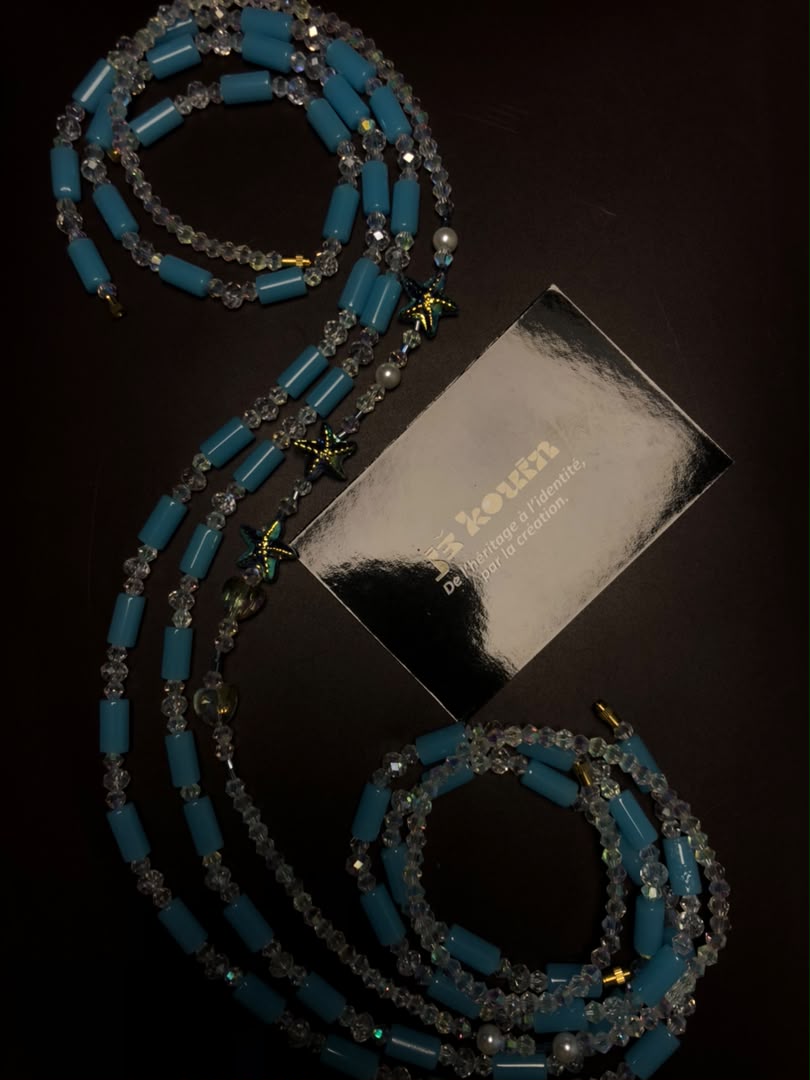 Chaîne de taille baya composée de tubes bleu turquoise et de perles de cristal transparentes ondulant sur fond sombre à côté d'une carte de visite jekouin argentée.